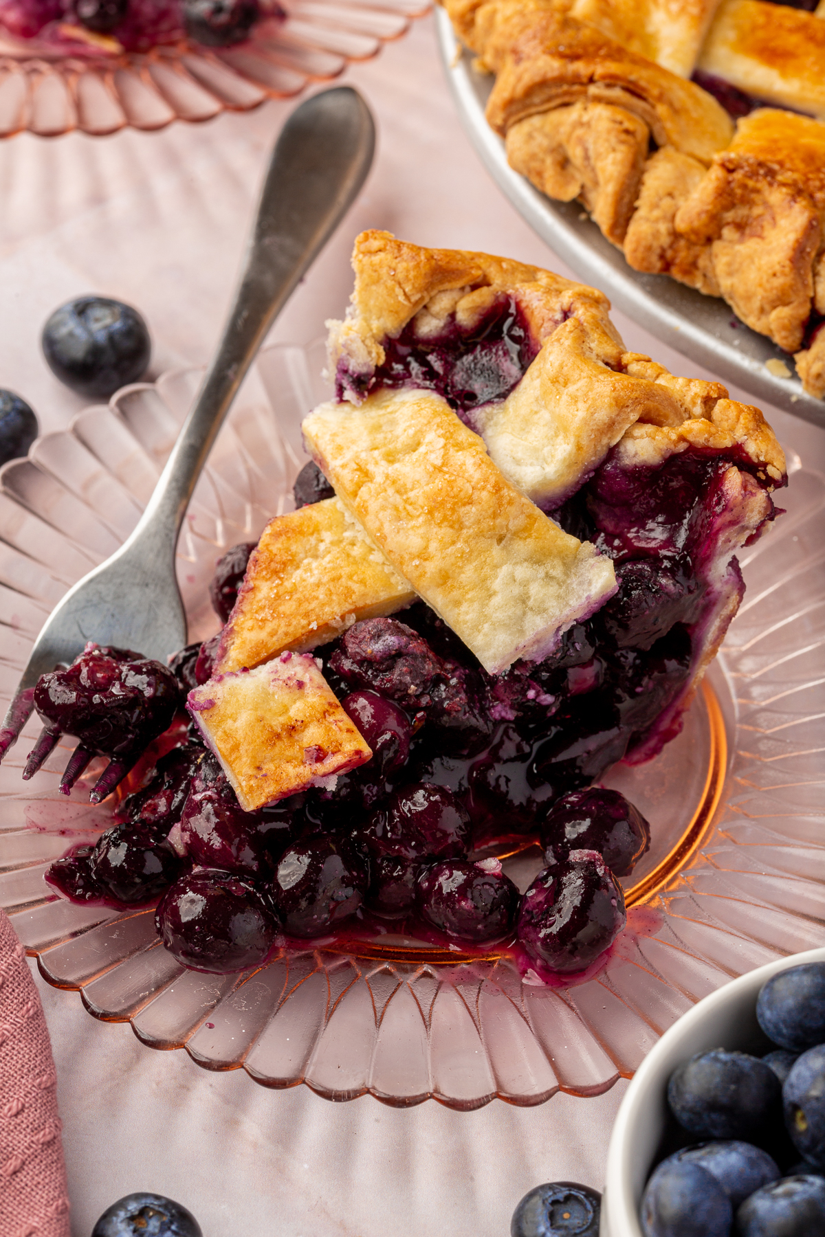 A slice of gluten-free blueberry pie on a glass plate with a fork cutting through the golden, flaky lattice crust, juicy blueberries spilling out. A whole pie is visible in the background, along with fresh blueberries scattered around.