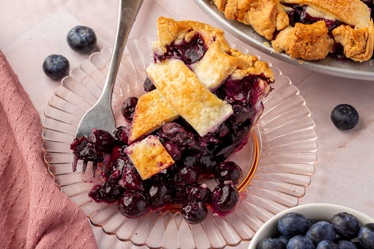 A horizontal view of a whole blueberry pie with several slices removed, showing the lattice crust and rich filling. Fresh blueberries are scattered around the scene.