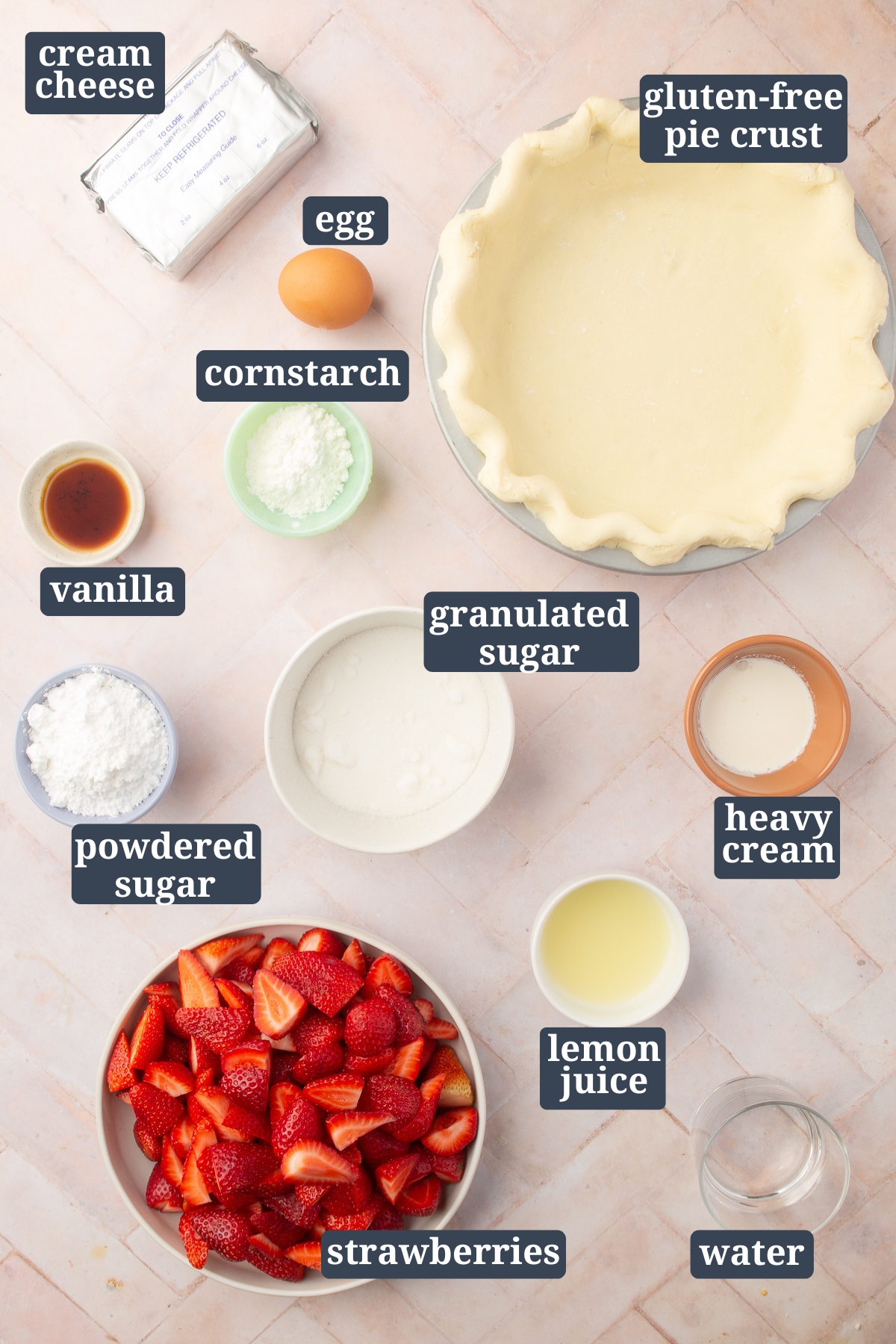 Overhead shot of gluten-free strawberry cream cheese pie ingredients laid out on a pink tiled surface, including labeled cream cheese, egg, cornstarch, gluten-free pie crust, vanilla, powdered sugar, granulated sugar, heavy cream, lemon juice, strawberries, and water.