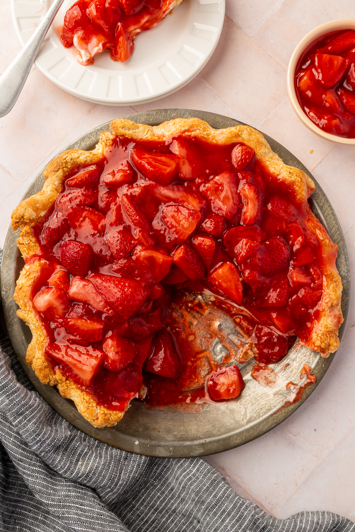 An overhead view of the strawberry cream cheese pie in a tin, with a slice missing and placed on a plate next to it. A small bowl of extra strawberry topping is off to the side.