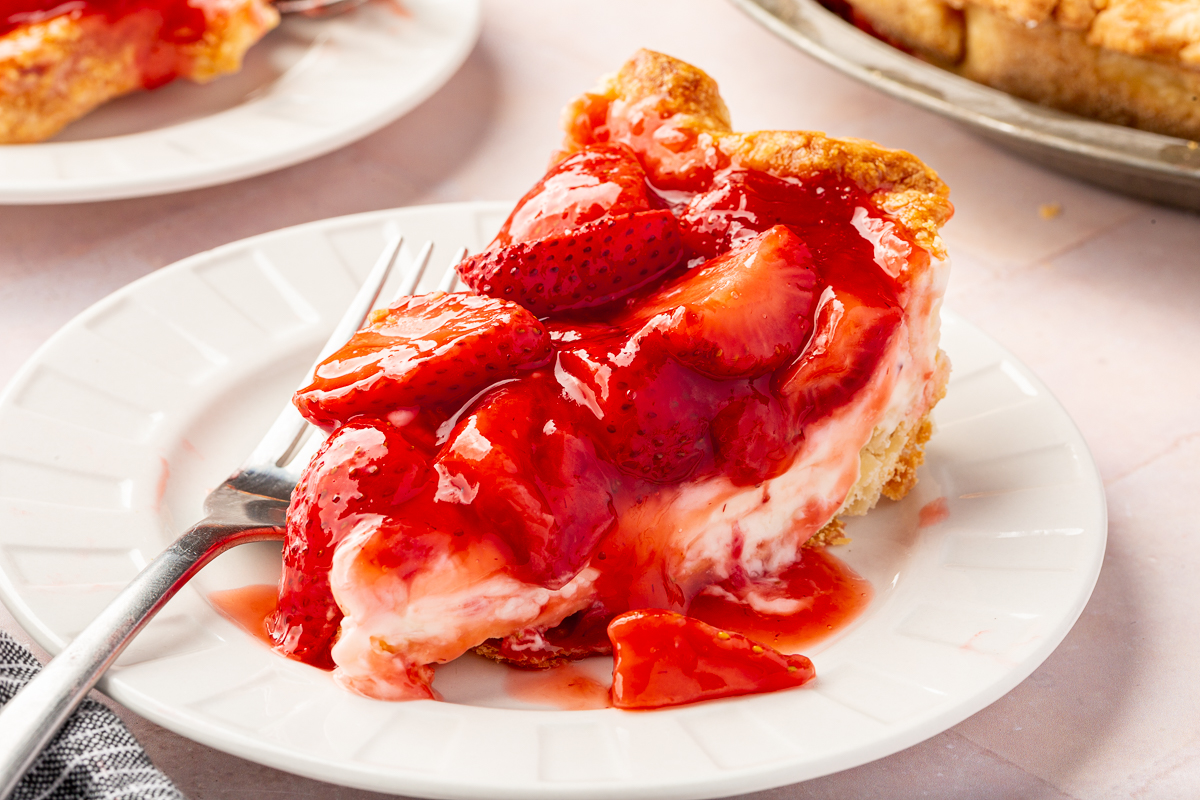 A single slice of gluten-free strawberry cream cheese pie sits on a white plate, topped with a glossy layer of fresh strawberries, with a fork placed beside it.