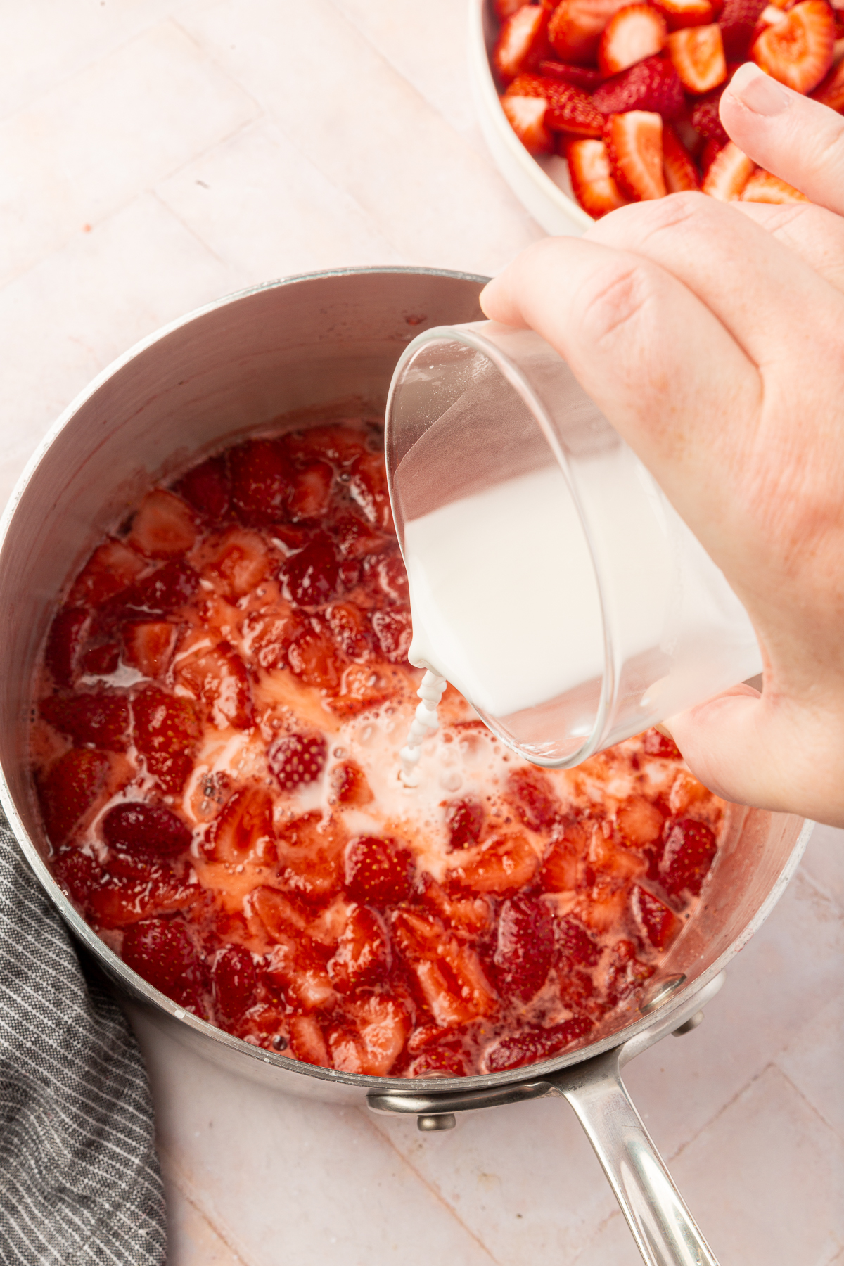 A hand pouring a white cornstarch slurry into a pot of simmering strawberries to thicken the mixture.