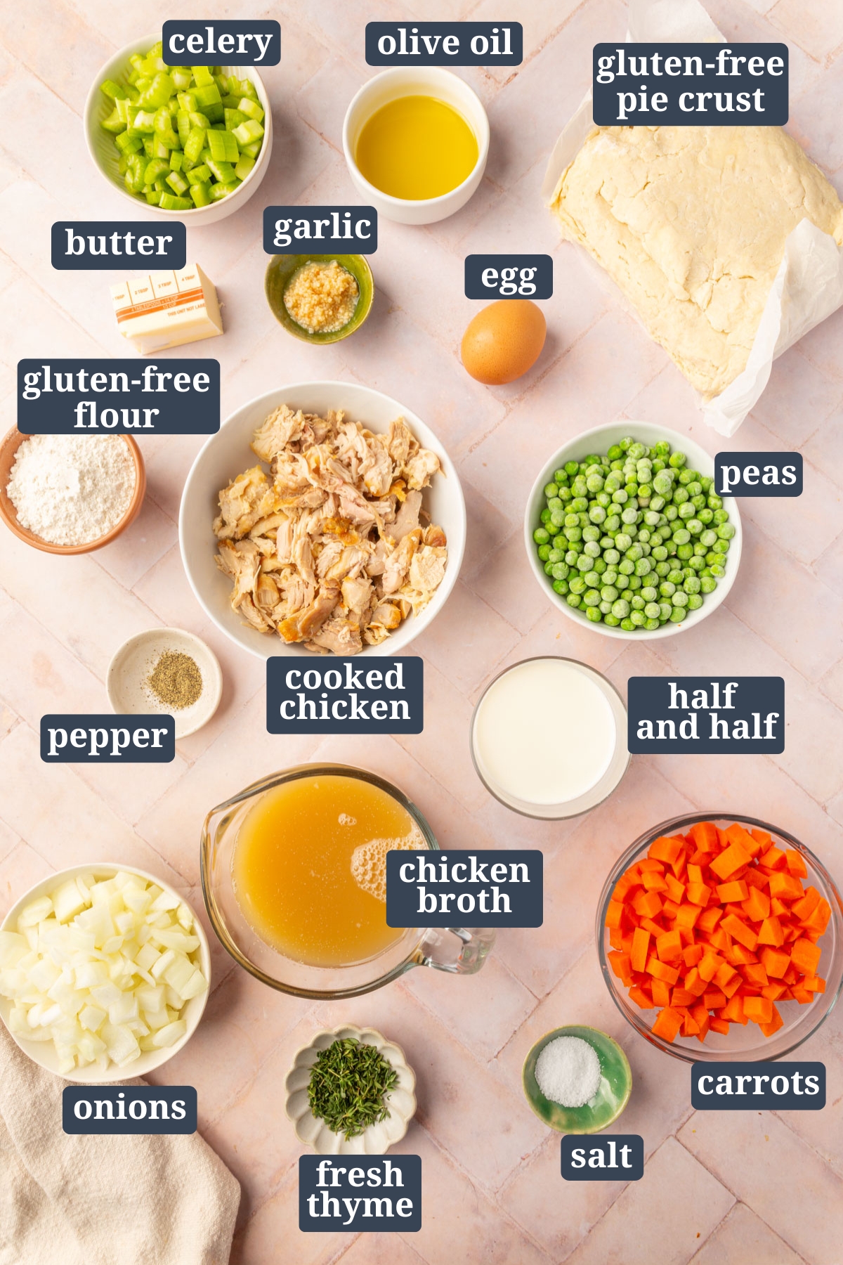 All ingredients for gluten-free chicken pot pie including celery, olive oil, gluten-free pie crust, butter, garlic, egg, gluten-free flour, cooked chicken, peas, pepper, half and half, chicken broth, onions, fresh thyme, salt and carrots all in individual bowls resting on a brick countertop.