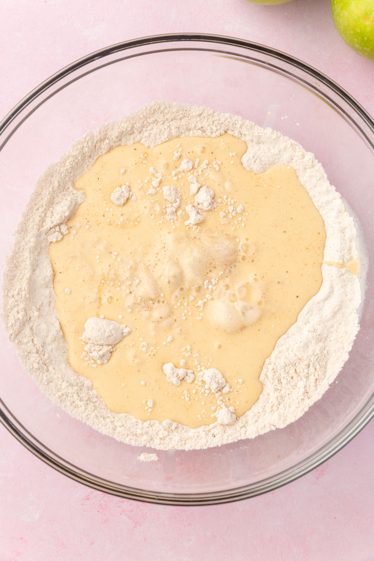 A glass mixing bowl of a wet mixture poured into the bowl of dry ingredients, ready to be whisked together into apple fritter batter.