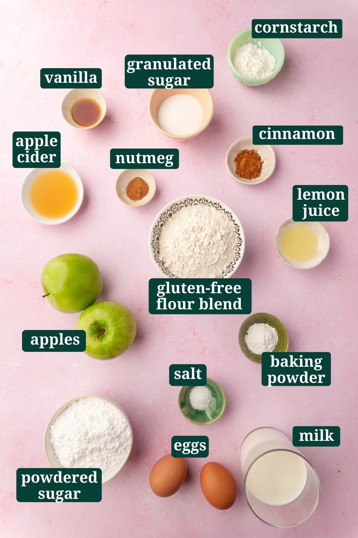 Overhead shot of gluten-free apple fritter ingredients laid out on a pink surface, including apples, gluten-free flour, sugar, cinnamon, and eggs, each labeled for easy identification.