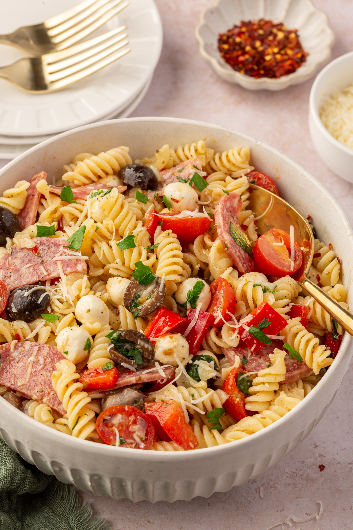 Photo of large serving bowl of gluten-free pasta salad with large serving spoon on table with various ingredients in the background.
