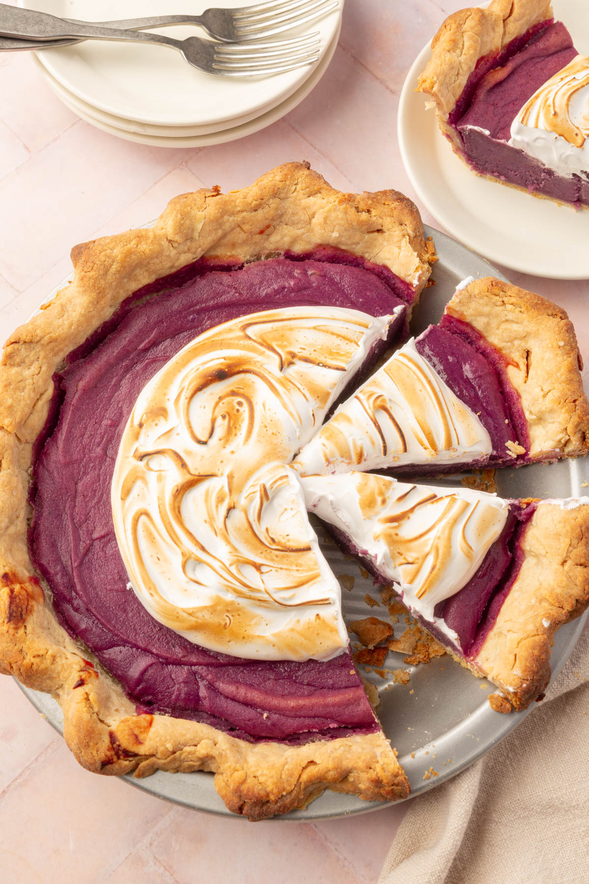 A close-up of a partially sliced baked purple sweet potato pie with toasted marshmallow meringue, a slice removed to reveal the vibrant purple filling and flaky crust.