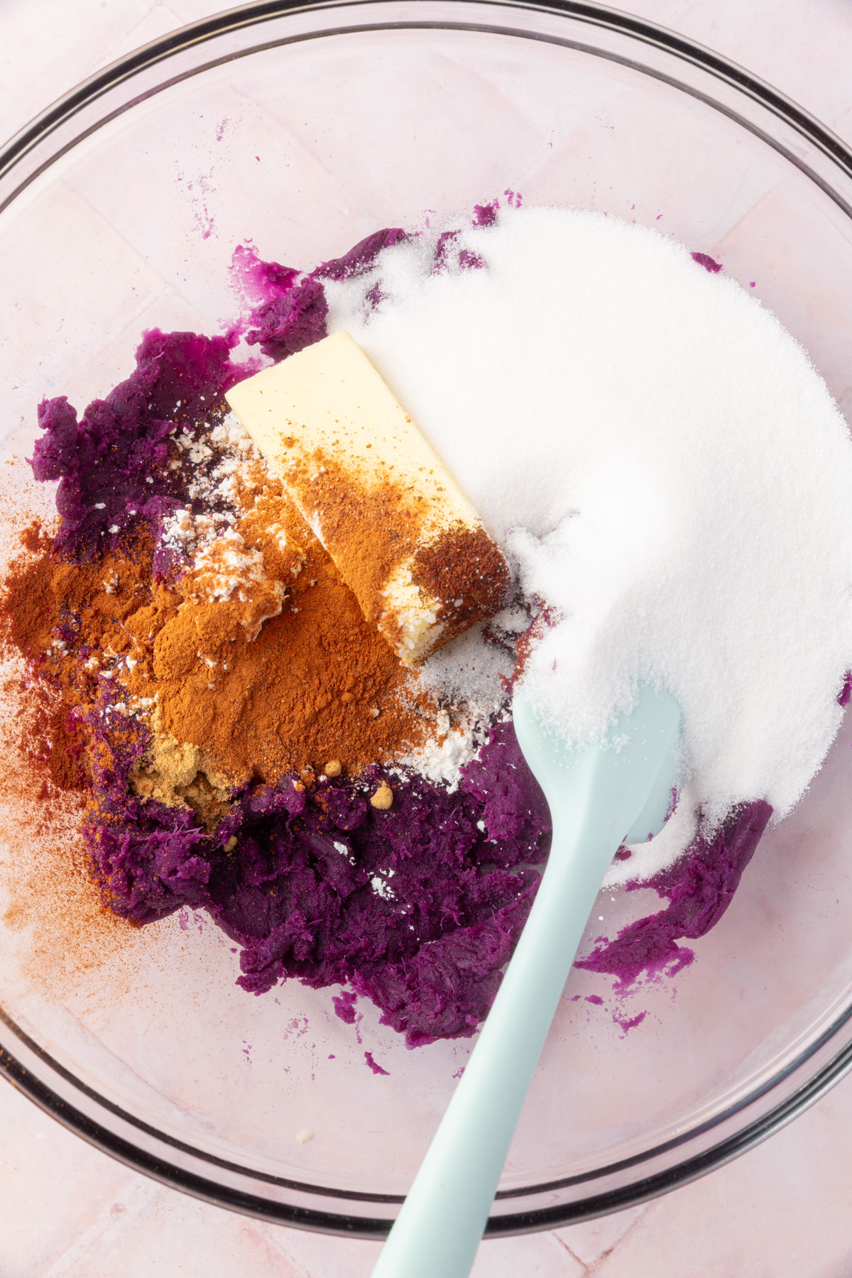 A bowl with mashed purple sweet potato, butter, granulated sugar, and spices like cinnamon and ginger, ready to be mixed for the pie filling.