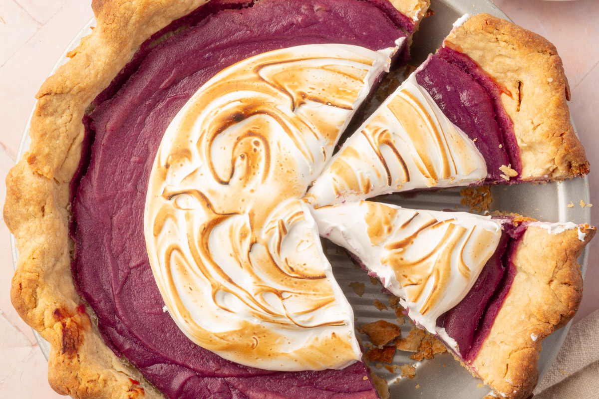 A full view of the partially sliced purple sweet potato pie with toasted meringue, styled with a stack of plates and forks in the background.