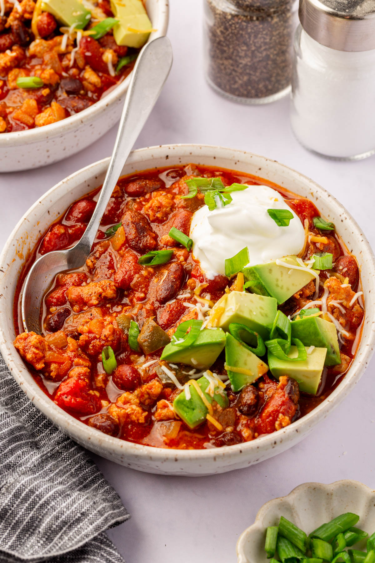 A bowl of slow cooker turkey chili topped with avocado, shredded cheese, sour cream, and chopped green onions, with a spoon inside. Another bowl, salt and pepper shakers, and a small dish of green onions are visible nearby.
