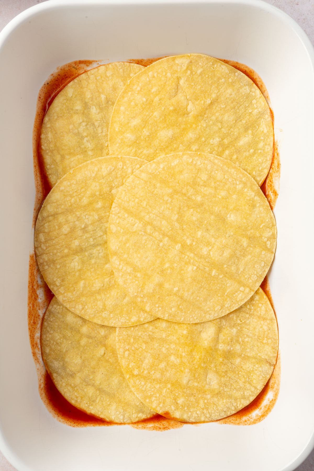 Large white baking pan with enchilada sauce layered on the bottom and corn tortillas on top.