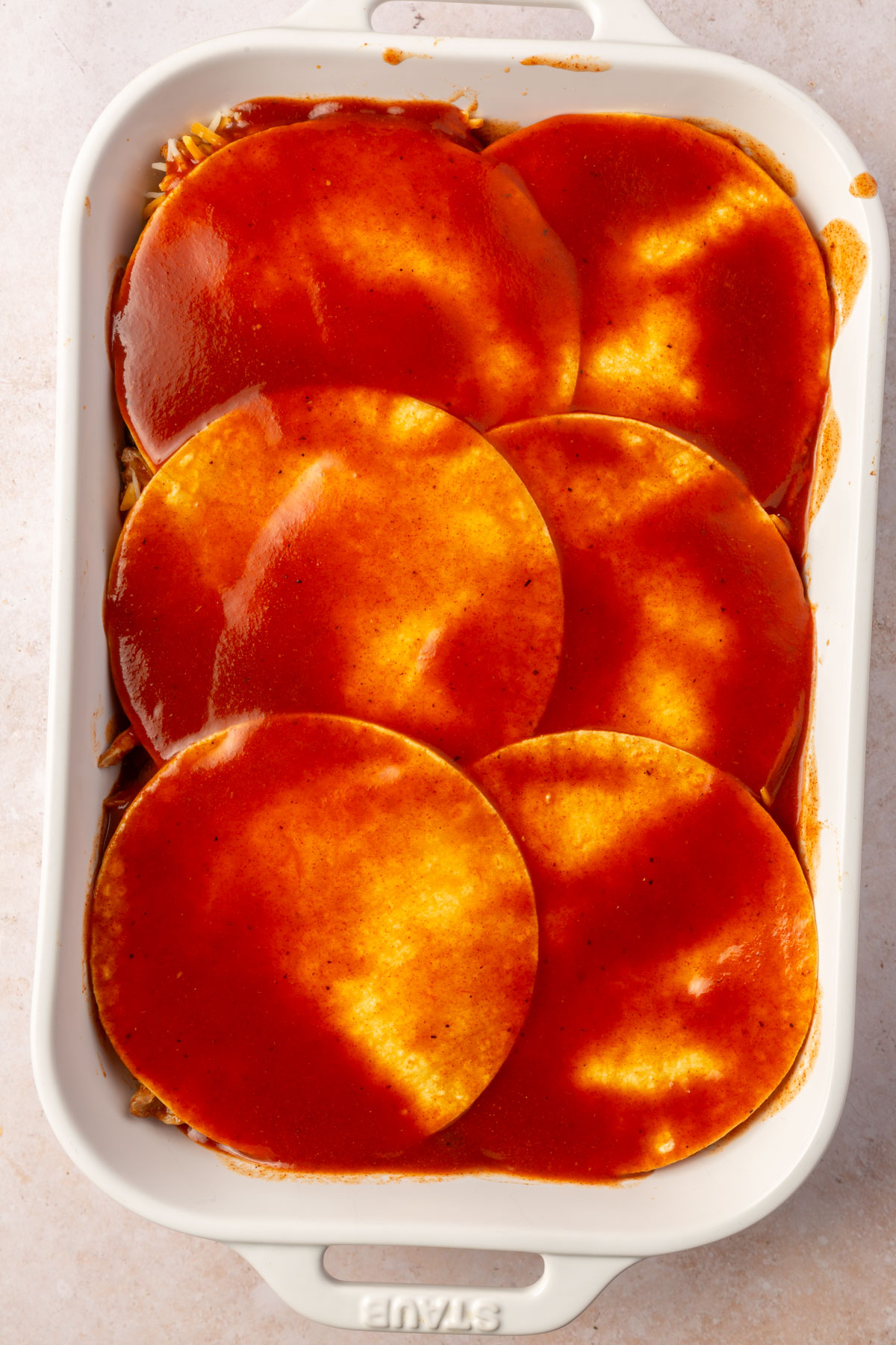 White baking pan with a layer of the chicken enchilada casserole already down and the corn tortillas covered with red enchilada sauce on top.