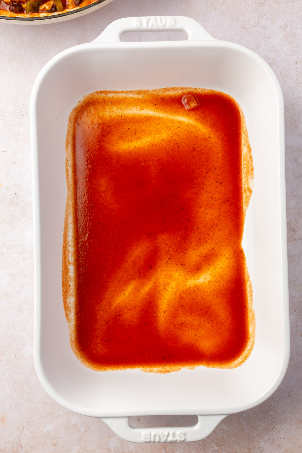 Large white baking pan with red enchilada sauce spread on the bottom of the pan.