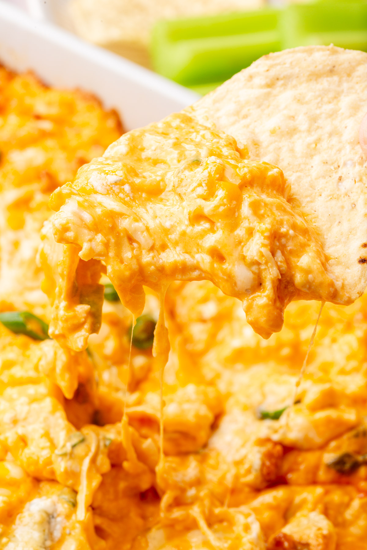 A tortilla chip lifting a scoop of buffalo chicken dip, with gooey melted cheese stretching from the dish.