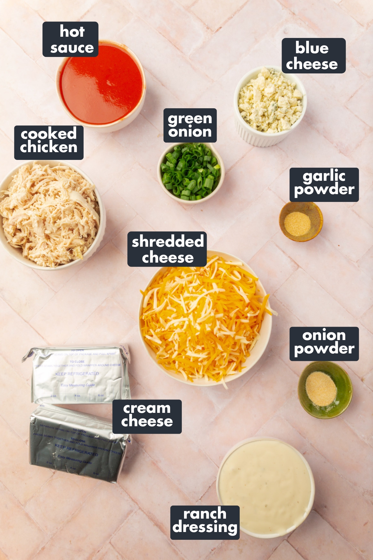 A flat lay of labeled ingredients for gluten-free buffalo chicken dip, including cooked chicken, shredded cheese, cream cheese, ranch dressing, hot sauce, blue cheese, green onions, garlic powder, and onion powder on a light pink tiled surface.