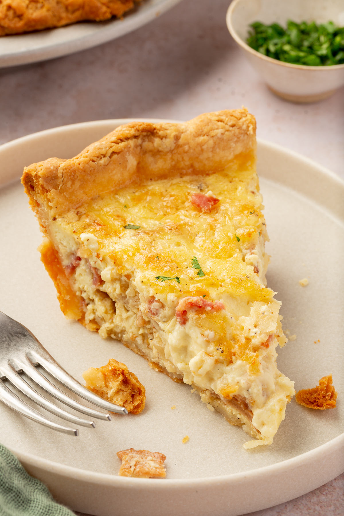 A slice of gluten-free quiche lorraine with bacon on a plate with a fork and few crumbs of the gluten-free pie crust on the plate.