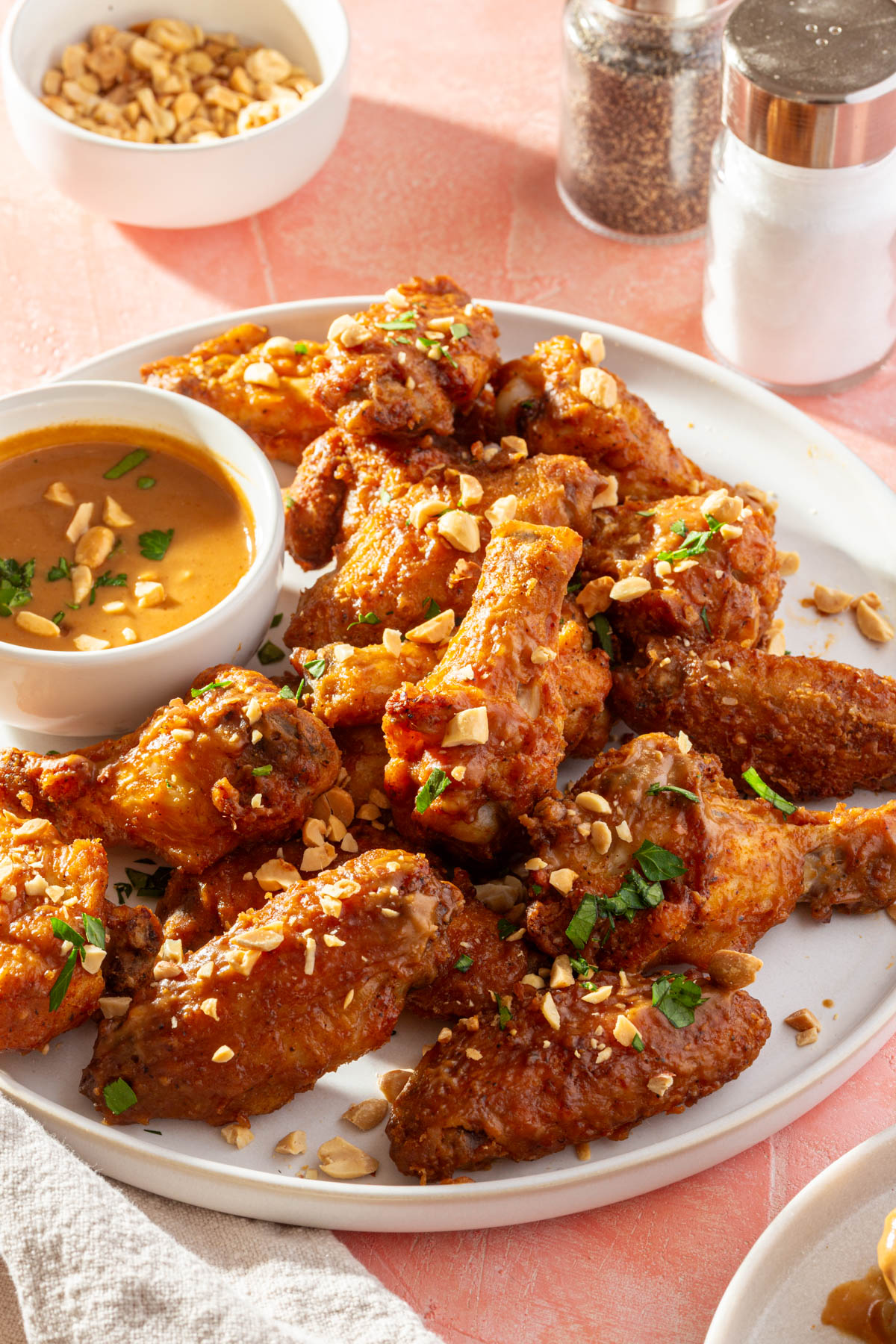 A plate of peanut butter chicken wings topped with crushed peanuts and parsley with a bowl of peanut butter sauce to the side, a salt and ppeper shaker and a bowl of chopped peanuts.