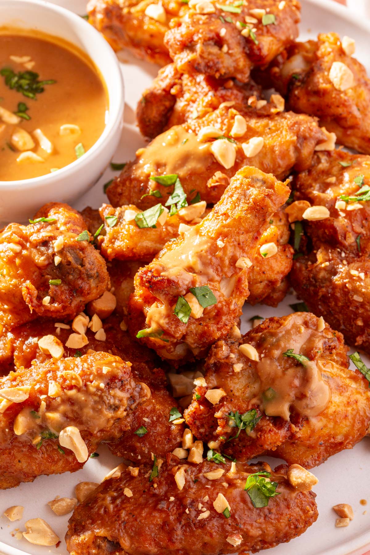 A close up of a plate of peanut butter chicken wings topped with chopped peanuts and parsley with a side of peanut butter sauce.