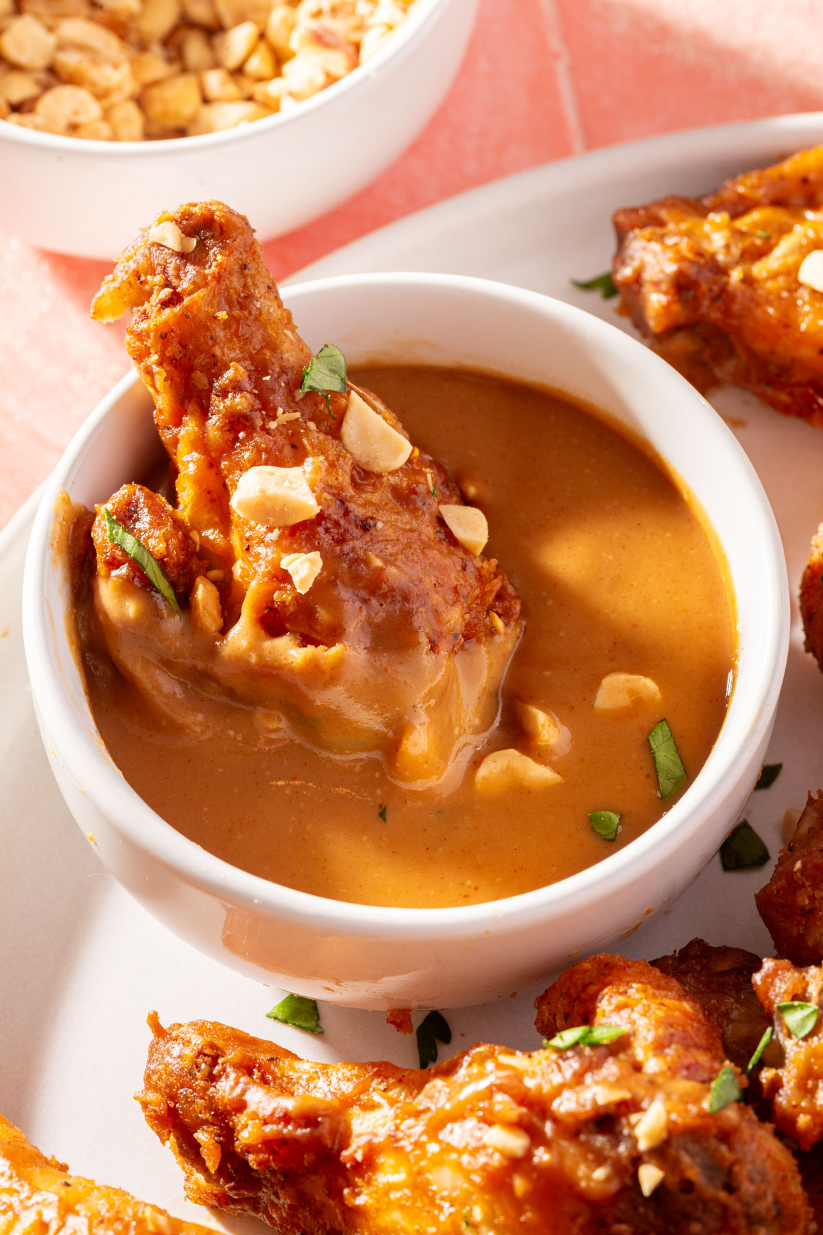 A peanut butter chicken wing being dipped in a ramekin of peanut butter sauce with a small bowl of chopped peanuts off to the side.