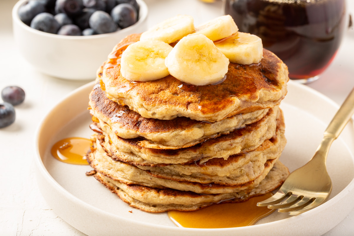 Stack of gluten-free banana pancakes on a white plate, topped with banana slices and drizzled with syrup, with a bowl of blueberries in the background.