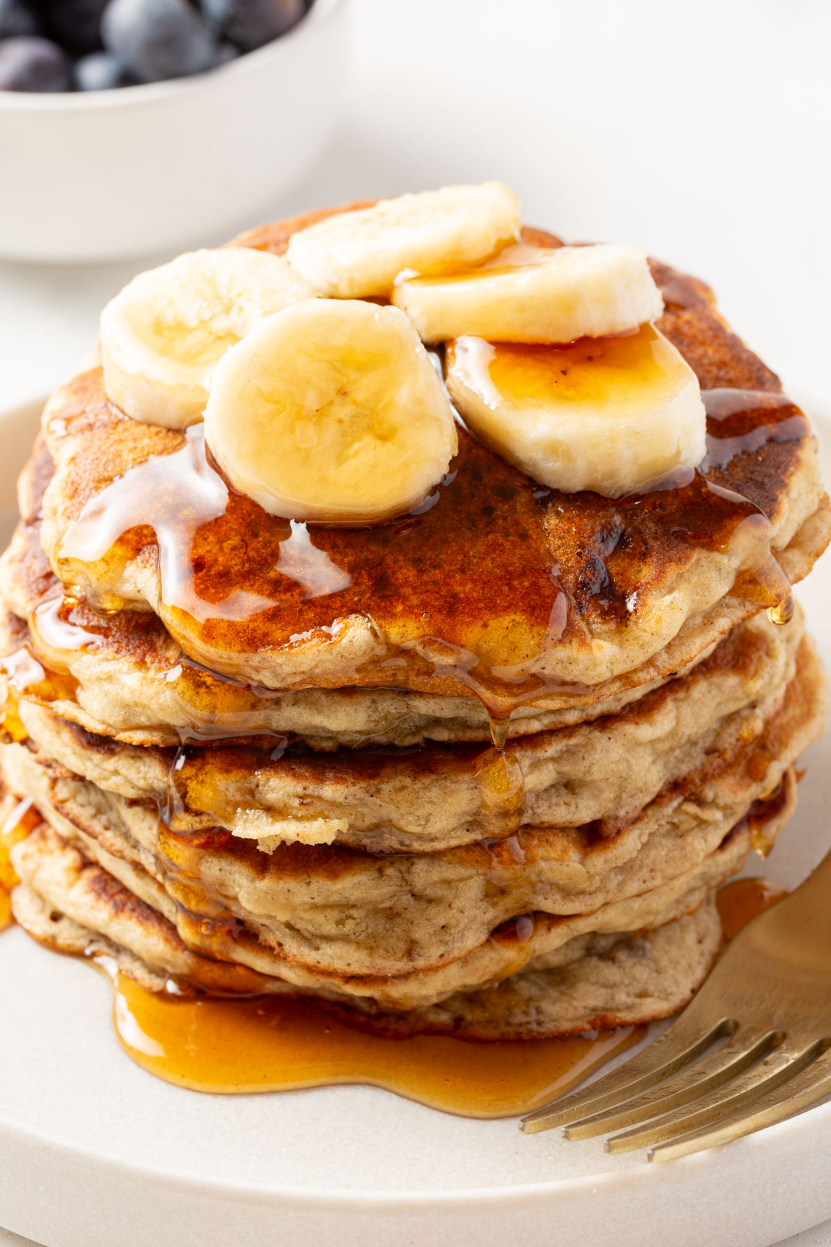 Gluten-free banana pancakes stacked on a plate, covered in syrup with banana slices on top, with a bowl of blueberries in the background.