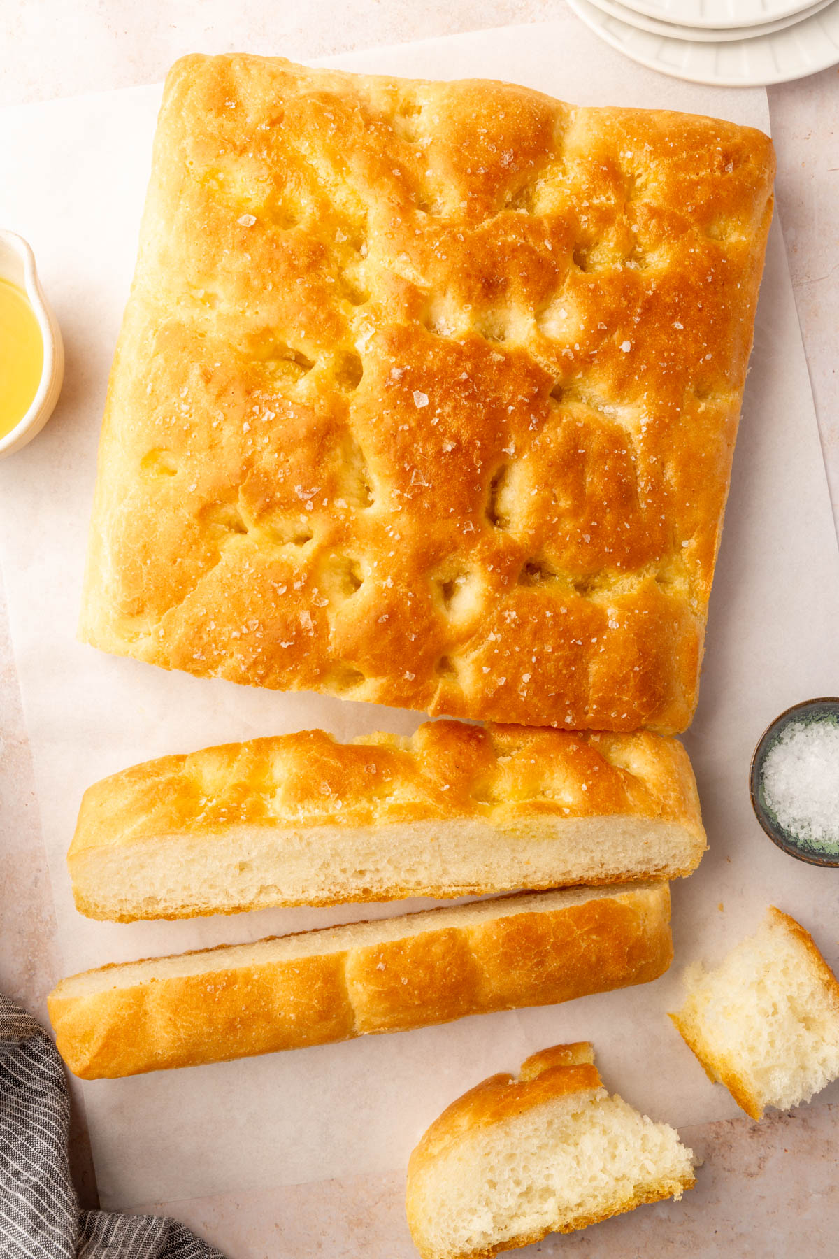 Overhead image of a full slab of baked gluten-free focaccia with a few slices cut to show texture, served on parchment with salt and oil on the side.