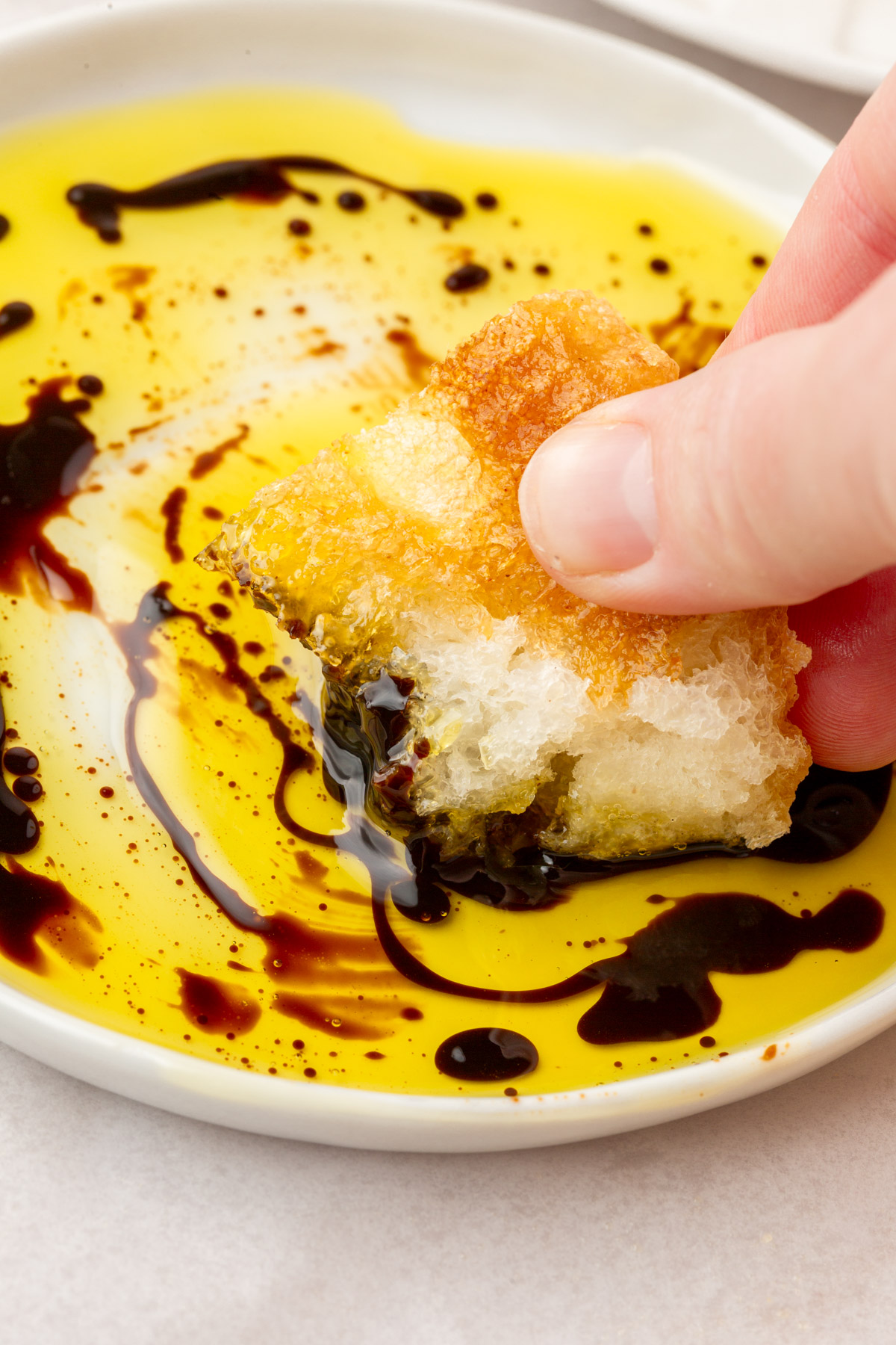 Hand dipping a torn piece of gluten-free focaccia into a swirl of olive oil and balsamic vinegar on a small white plate.