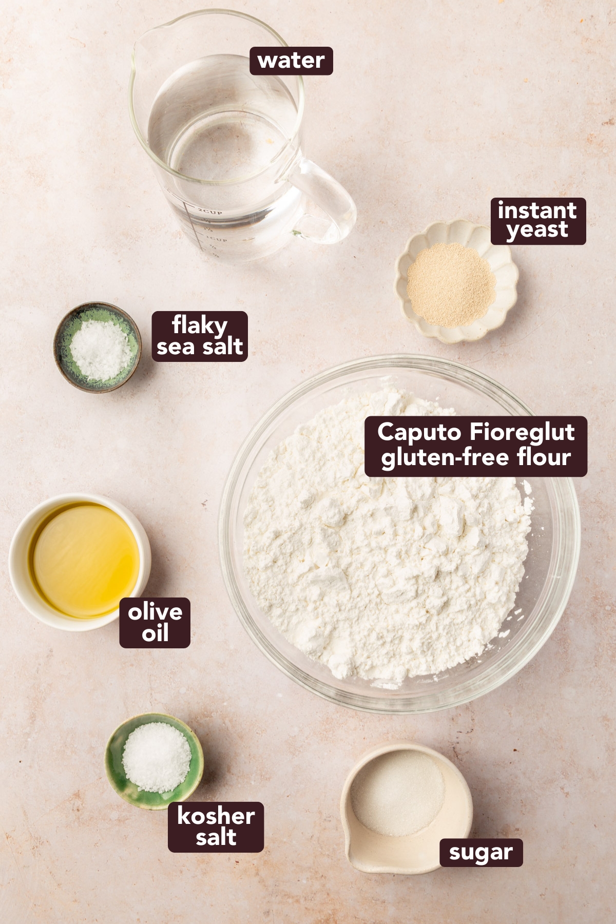 Overhead shot of ingredients for gluten-free focaccia, including Caputo Fioreglut flour, instant yeast, warm water, olive oil, sugar, kosher salt, and flaky sea salt.