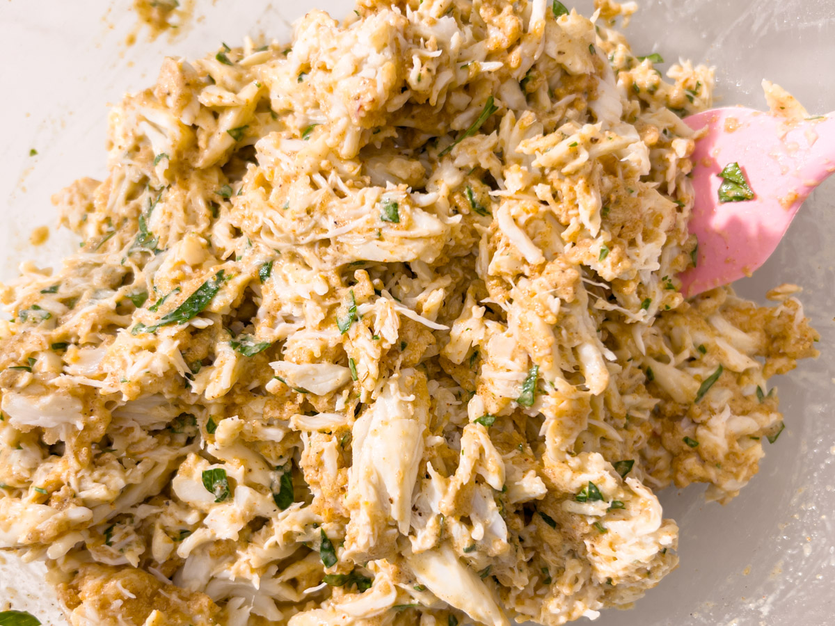 Fully mixed gluten-free crab cake filling with parsley and crab, being folded together with a pink spatula in a glass bowl.