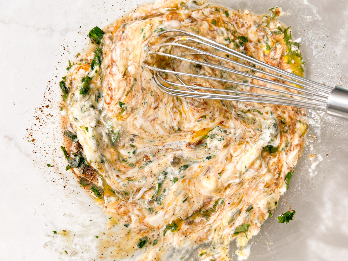 Whisk mixing creamy crab cake base with herbs and spices in a glass bowl.