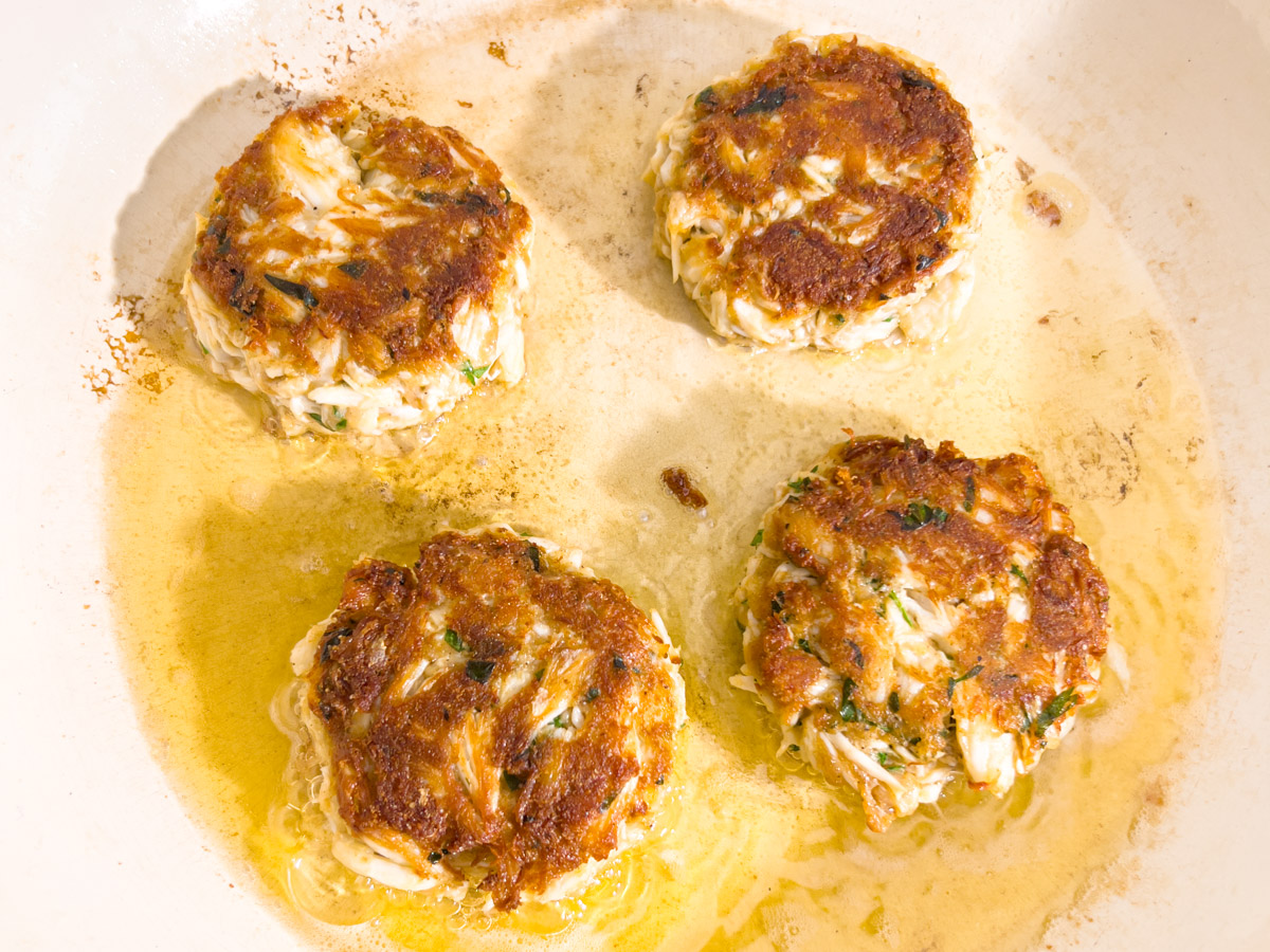 Gluten-free crab cakes frying in oil with golden brown crust forming.