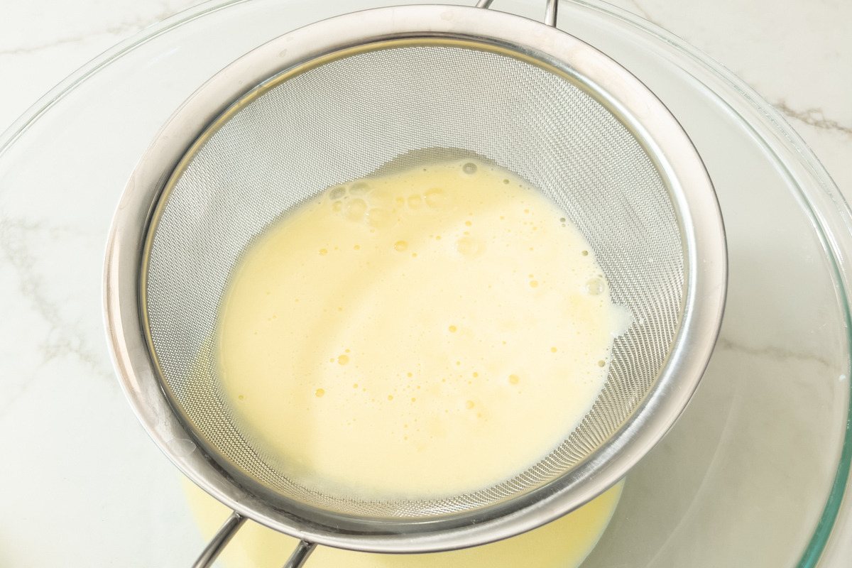 Ice cream custard being strained through a fine mesh sieve into a glass bowl to remove lumps.