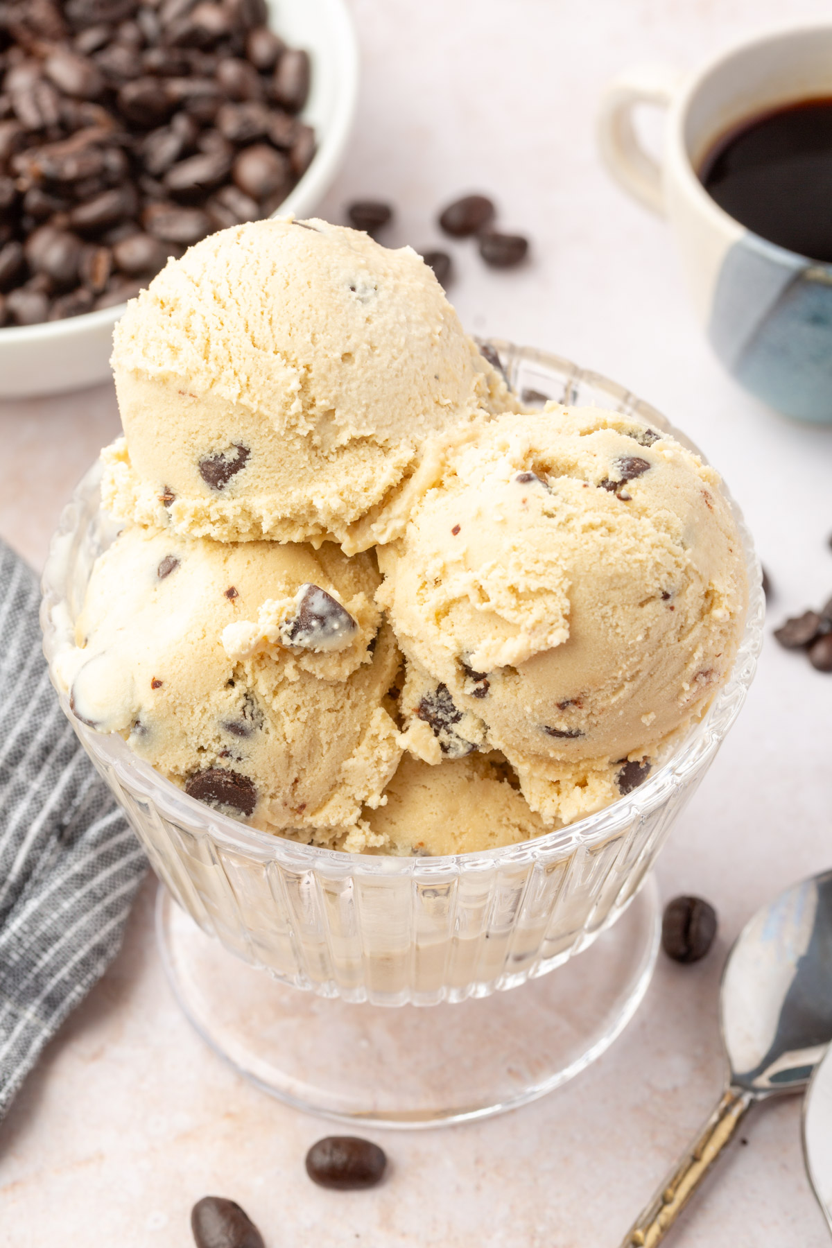 Scoops of espresso chocolate chip ice cream in a fluted glass dish, surrounded by coffee beans and a striped cloth napkin.