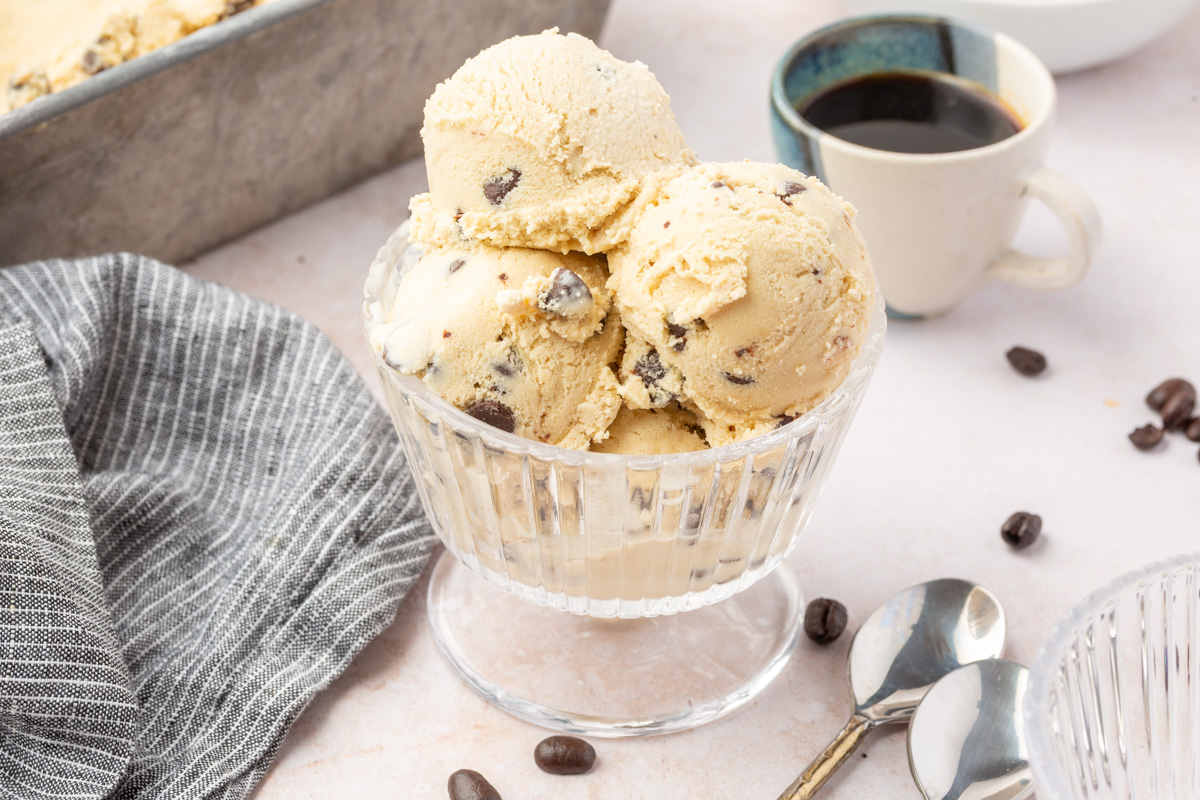 Espresso chocolate chip ice cream served in a glass bowl next to cups of coffee and metal spoons on a light background.