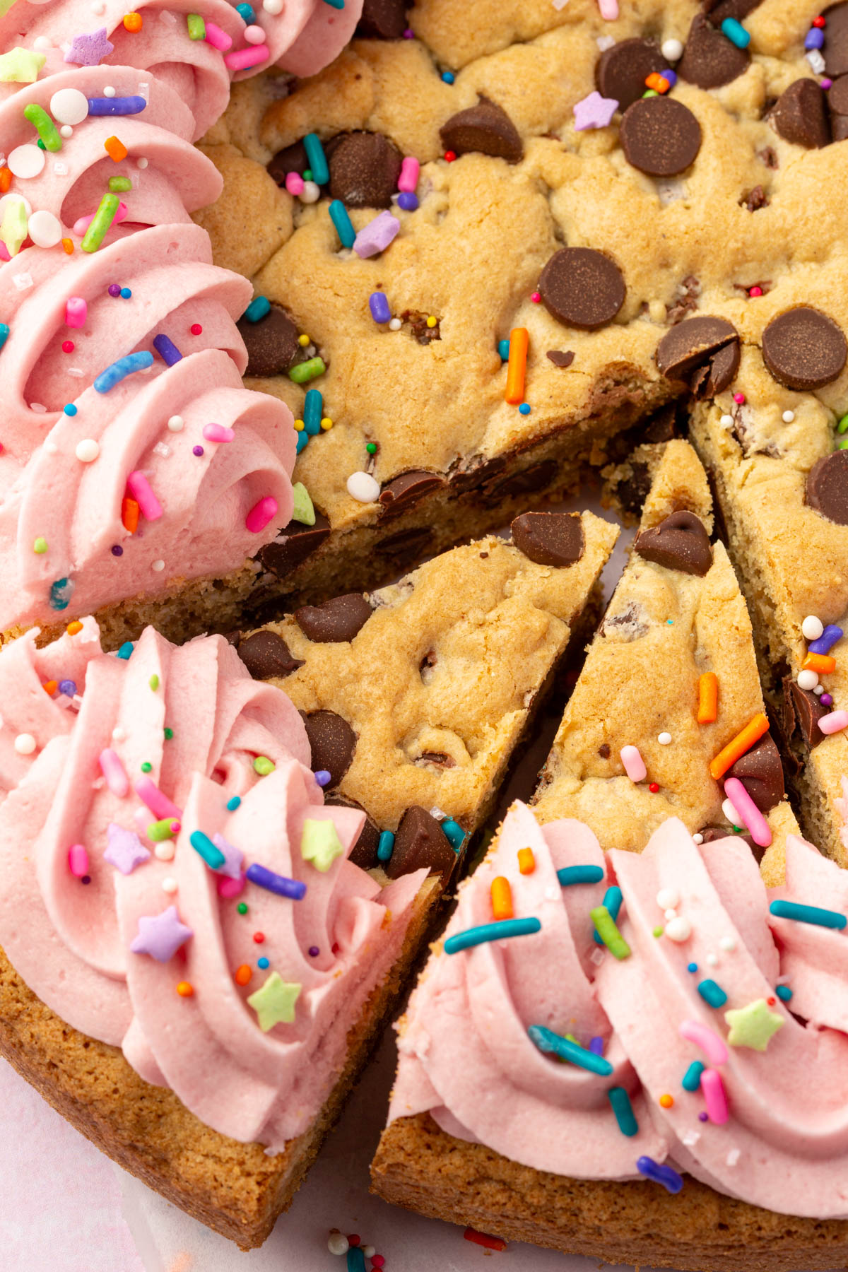 Close-up view of two slices cut from a gluten-free cookie cake, showing chocolate chips and pink piped frosting topped with sprinkles.