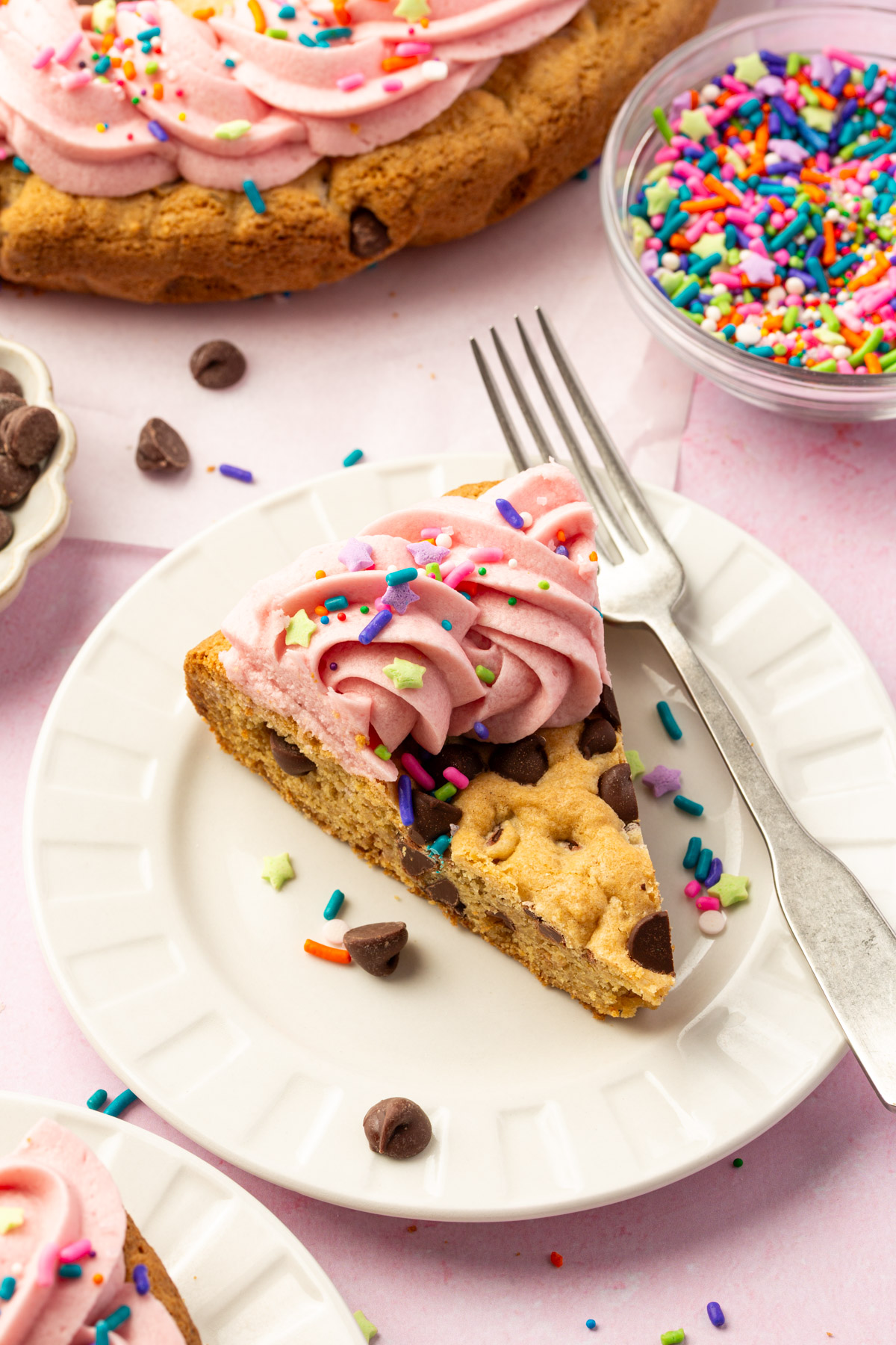 Slice of gluten-free cookie cake on a white plate, topped with pink buttercream frosting and sprinkles, surrounded by bowls of chocolate chips and sprinkles.