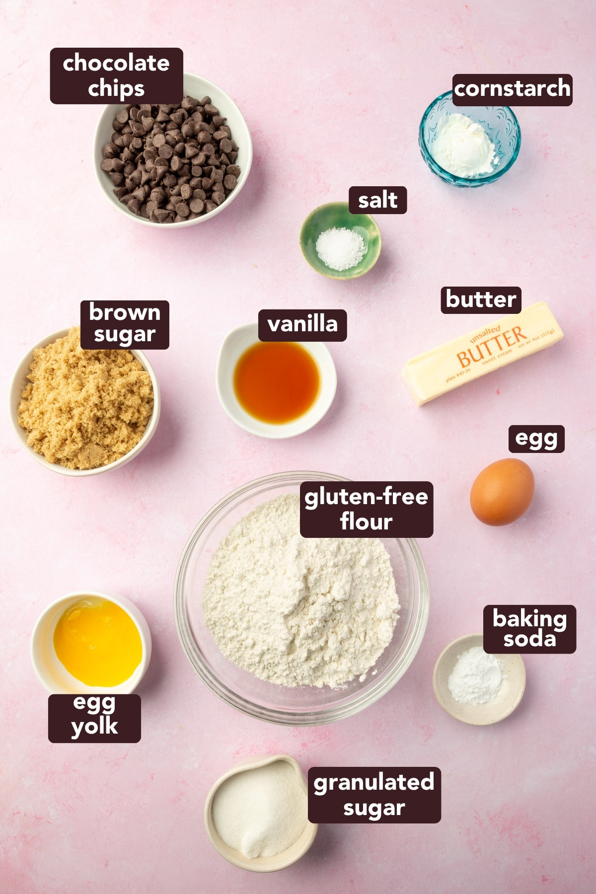 Overhead of labeled ingredients for a gluten-free cookie cake, including gluten-free flour, butter, sugars, vanilla, cornstarch, and chocolate chips on a pink background.