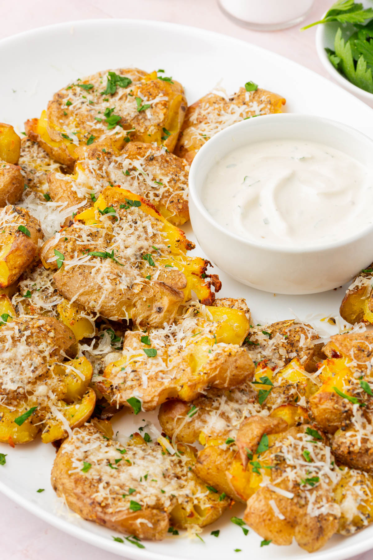Crispy parmesan smashed potatoes served on a white platter with a bowl of creamy dipping sauce.