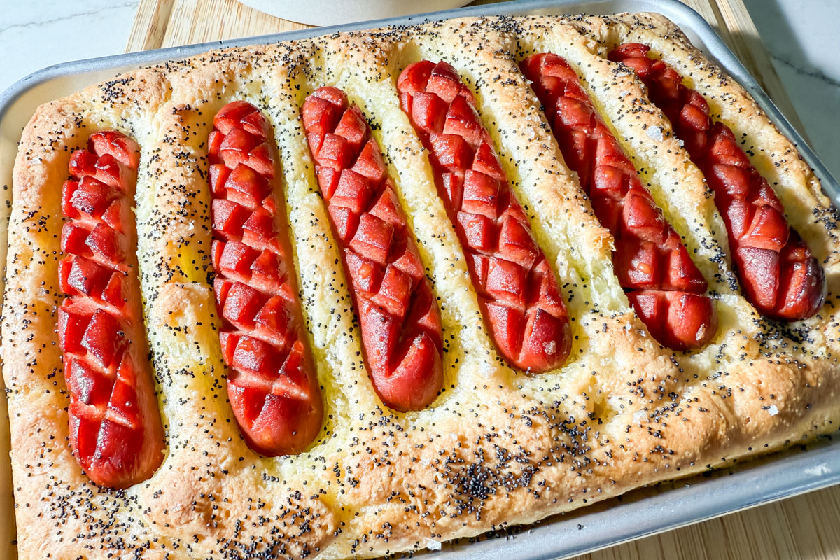 Close-up of golden-brown gluten-free hot dog focaccia with crispy, crosshatched hot dogs nestled in olive oil–rich bread.