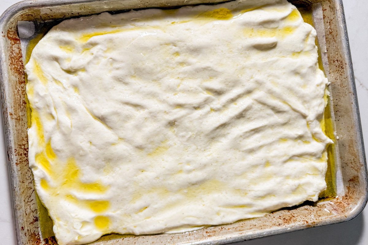 Gluten-free focaccia dough spread into an oiled sheet pan, ready for dimpling.