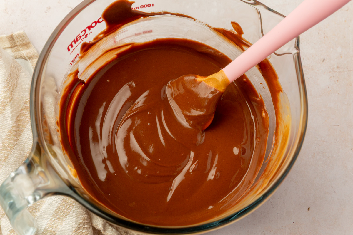A glass bowl filled with smooth, glossy melted chocolate and peanut butter mixture, stirred with a pink spatula.