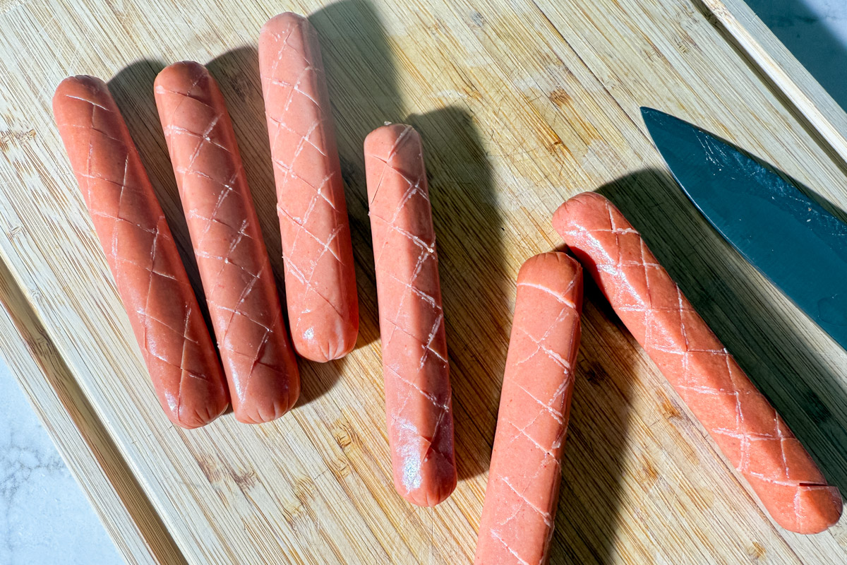 Raw hot dogs crosshatched with a knife on a wooden cutting board, ready to be baked into focaccia.