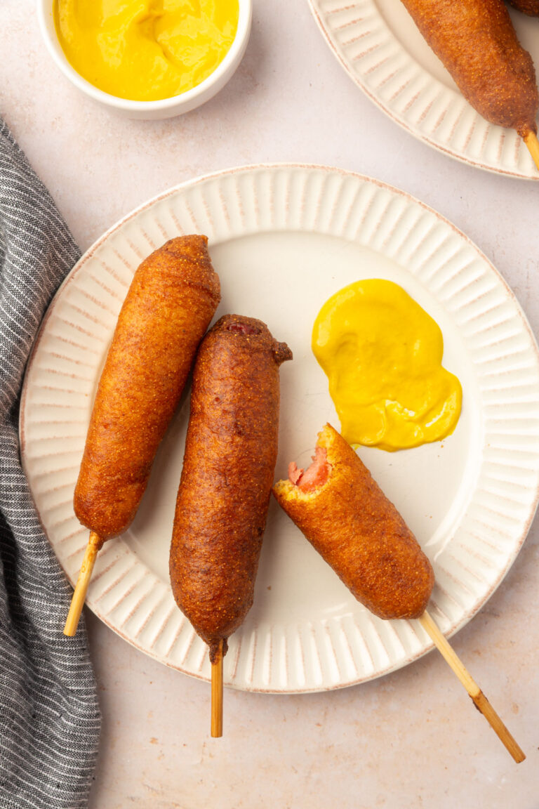 Three gluten-free corn dogs on a white plate served with yellow mustard for dipping.