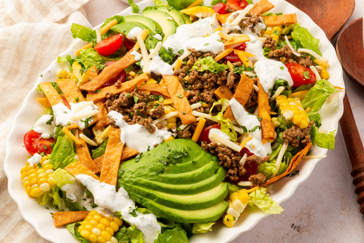 Gluten-free taco salad with homemade fried tortilla strips, avocado slices, corn kernels, ground beef, and creamy dressing.