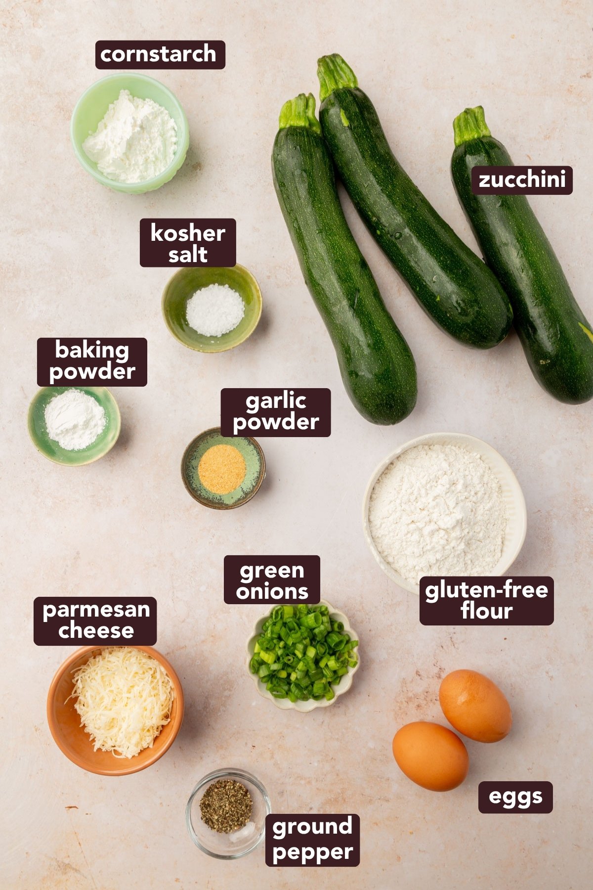 Overhead view of ingredients for gluten-free zucchini fritters, including fresh zucchini, gluten-free flour, green onions, eggs, parmesan, and spices.