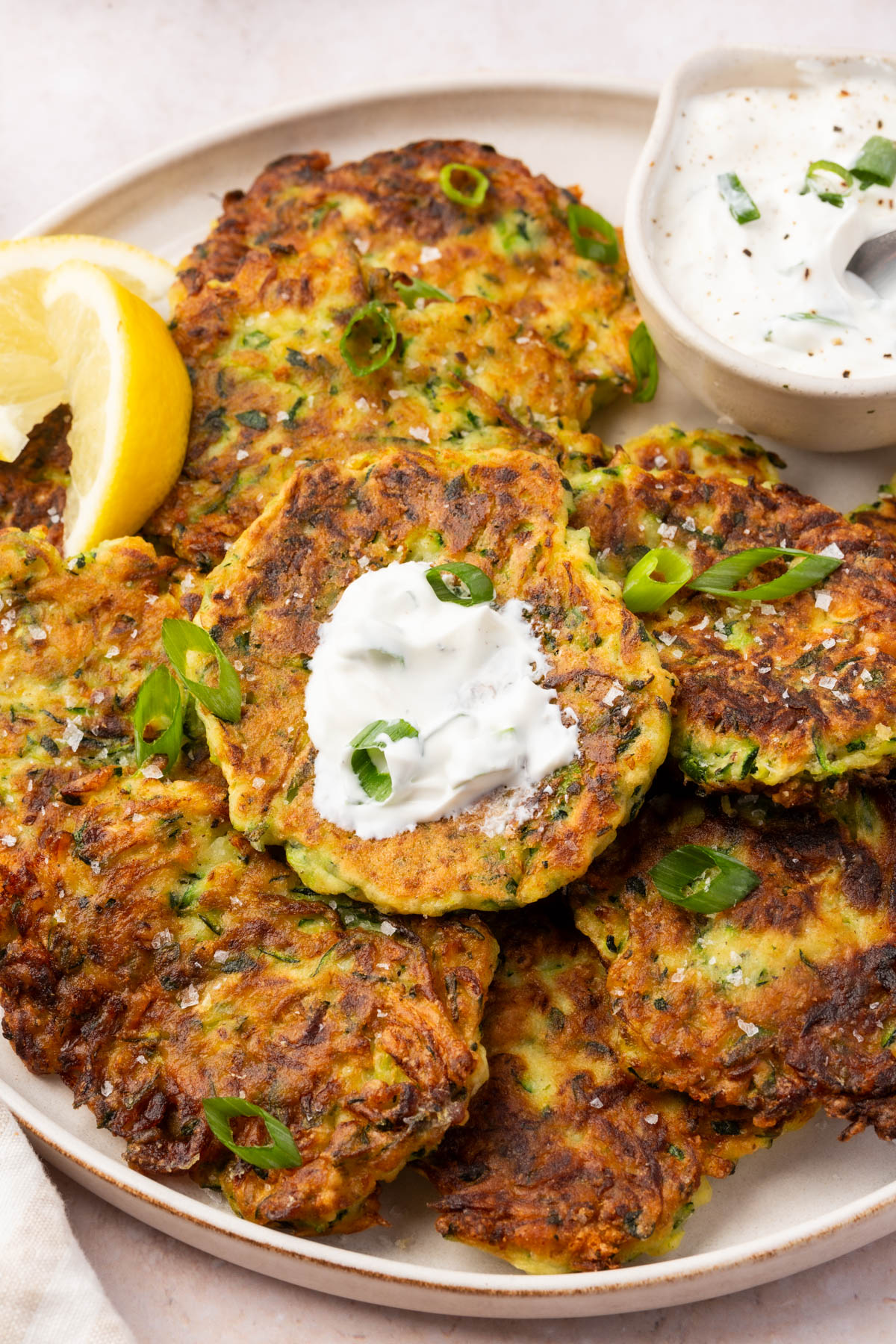 Gluten-free zucchini fritters topped with a dollop of sour cream and garnished with green onion, served with lemon slices and creamy dip on the side.