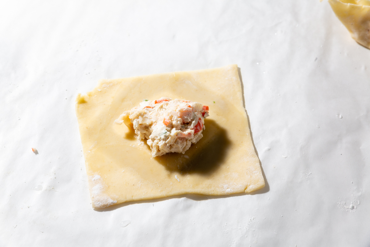 A square gluten-free wonton wrapper topped with a scoop of creamy crab rangoon filling and a diamond of water surrounding the filling.
