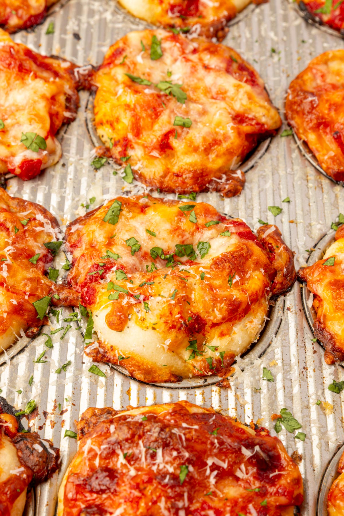 Baked gluten-free focaccia pizza muffins in a muffin tin topped with melted mozzarella, marinara, and herbs.