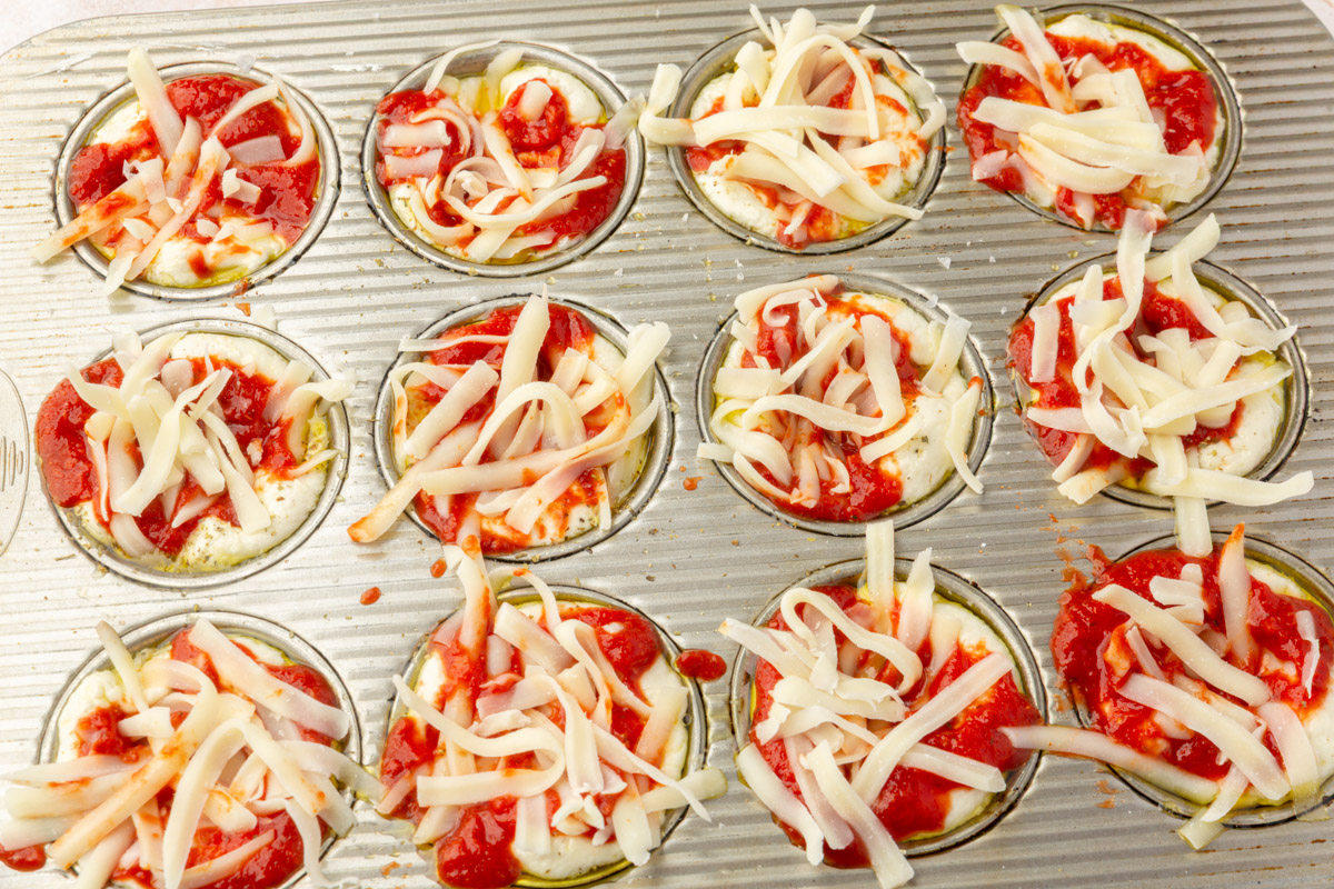 Muffin tin filled with focaccia dough topped with pizza sauce and shredded mozzarella cheese before baking.