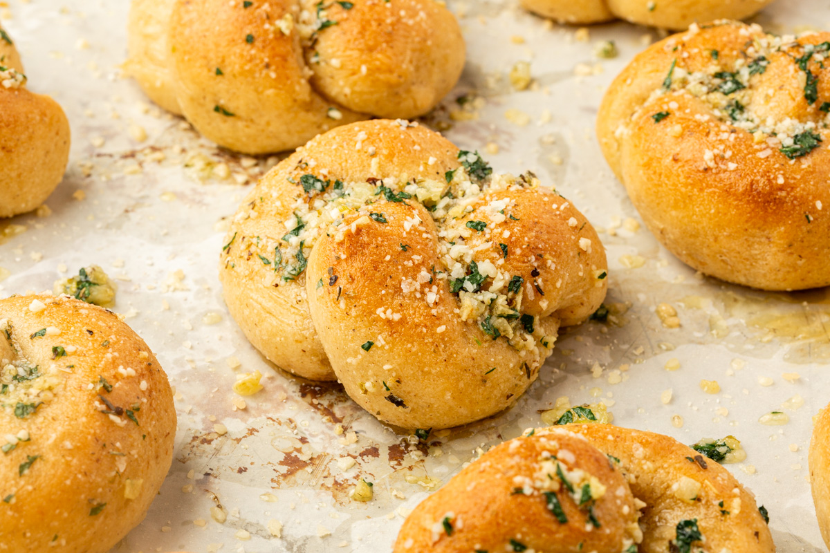 Gluten-free garlic knots on a parchment-lined baking sheet, brushed with garlic butter and sprinkled with parsley and cheese.